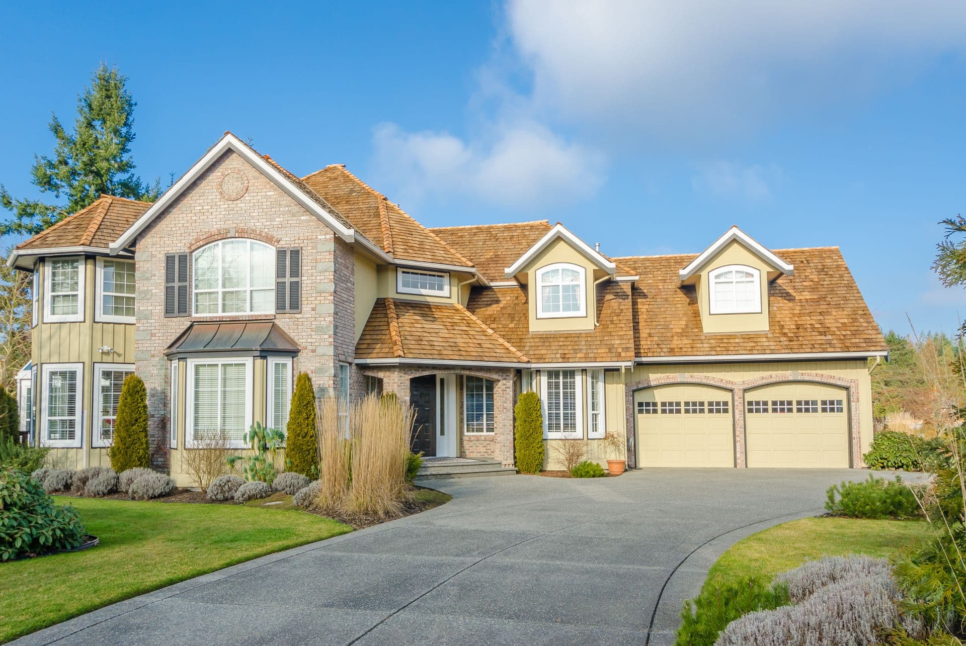 A large, elegant suburban house with a brick facade, multiple gable roofs, and large windows. A well-manicured lawn lines the curved driveway.