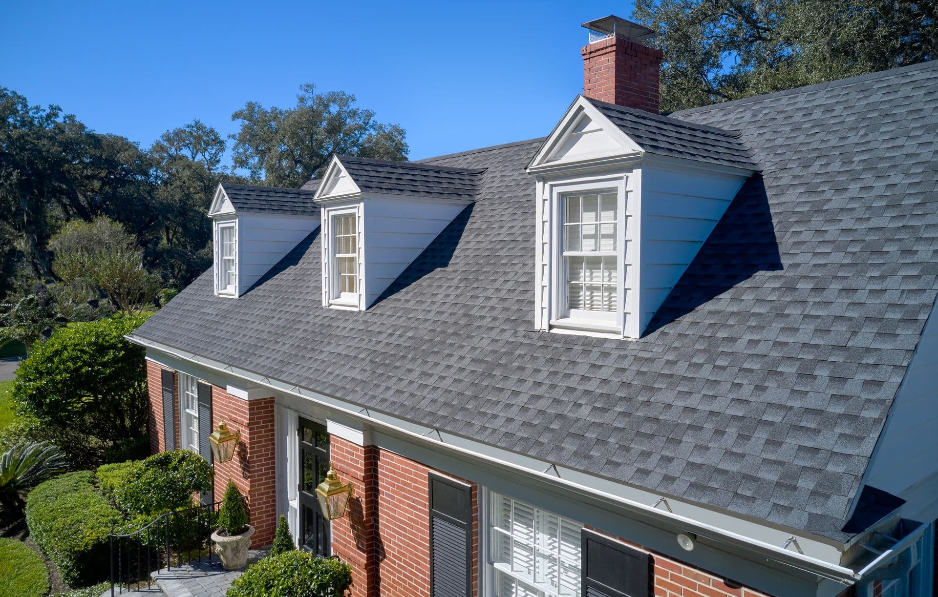 A brick house with a dark shingle roof, featuring three dormer windows. Lush green bushes line the front, enhanced by clear blue sky backdrop.