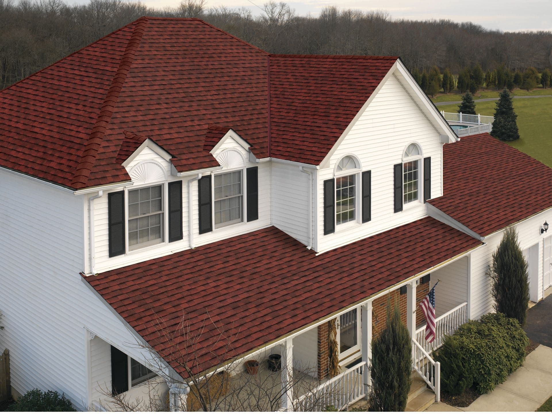 A two-story white house with red gable roof shingles, black shutters, and arched windows. A porch displays an American flag, set in a wooded area.