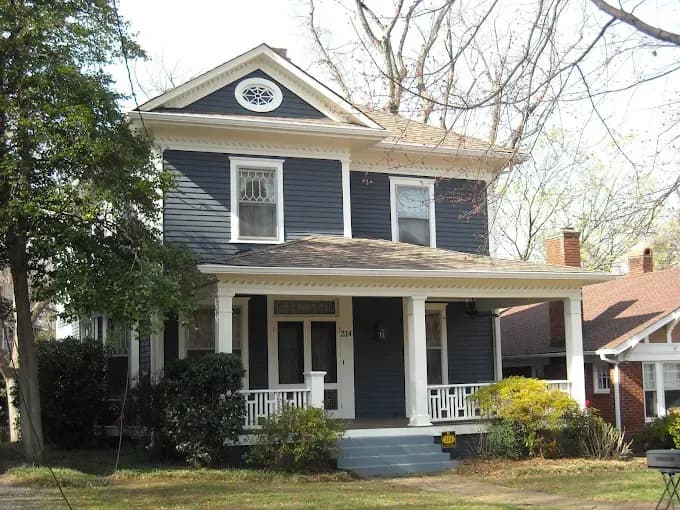 A charming two-story house with dark blue siding and white trim, featuring a covered porch, surrounded by bare trees and shrubs, exuding a cozy feel.